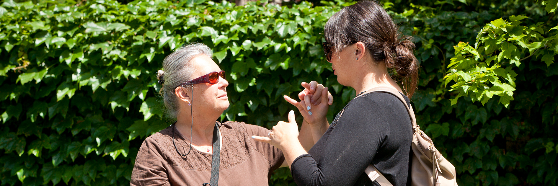Une accompagnatrice et une cliente se tiennent dehors par une journée ensoleillée et utilisent l’ASL tactile pour communiquer.