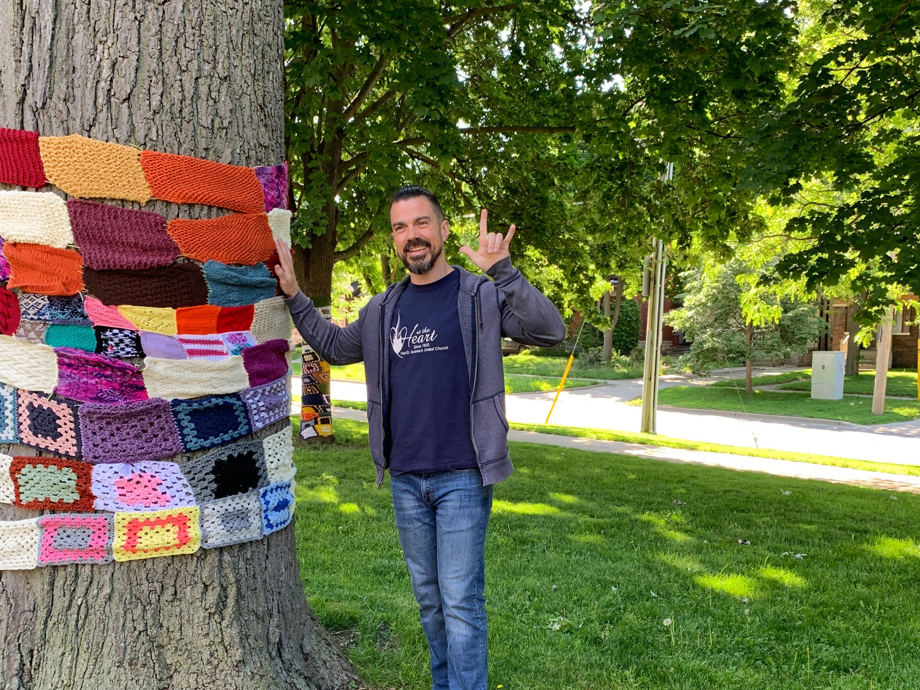 A DBCS staff member displays the sign in ASL for I love you while standing beside a tree that has been yarn bombed
