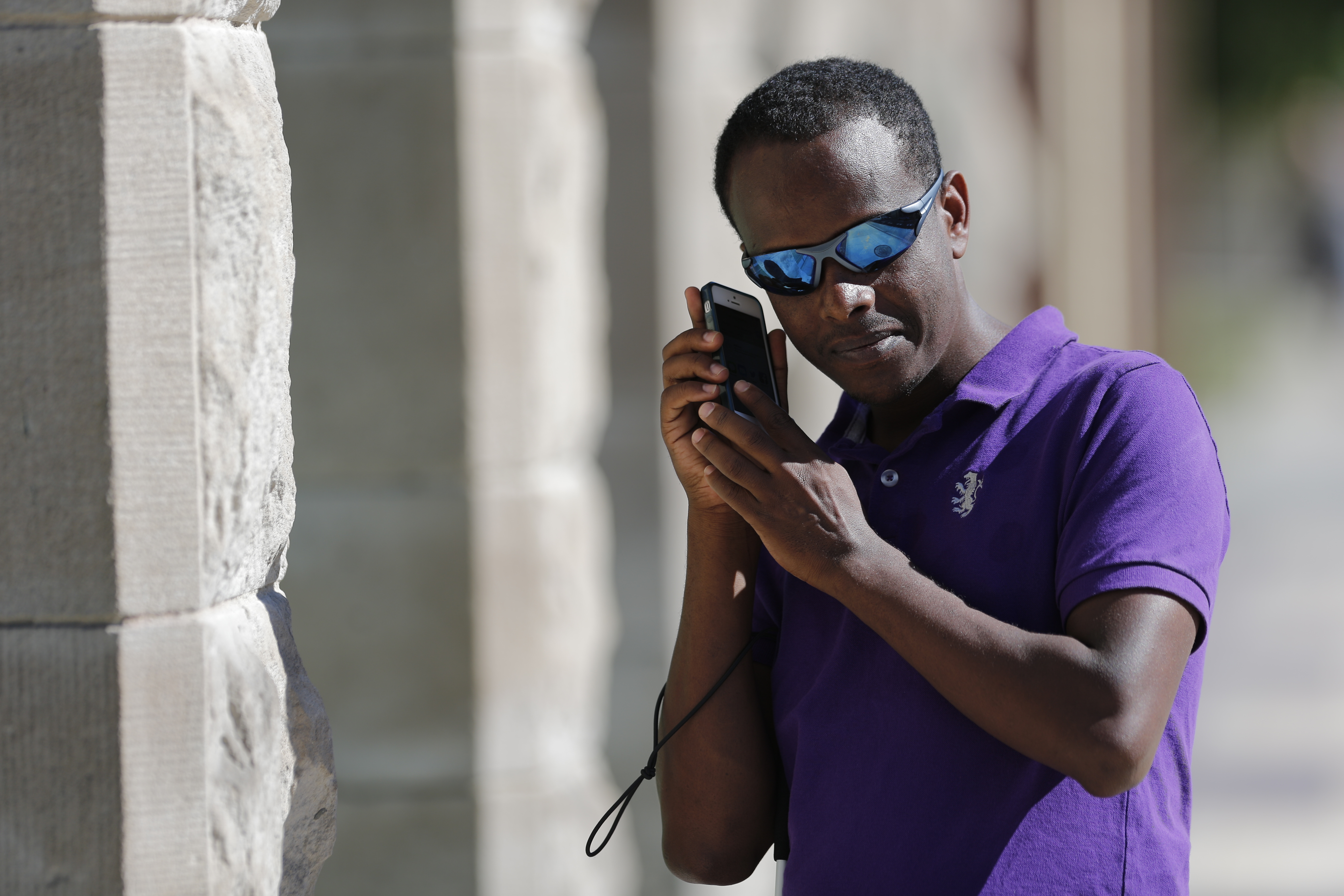 Un homme portant des lunettes de soleil porte un smartphone à son oreille pour écouter. Il tient une canne blanche sous son bras.