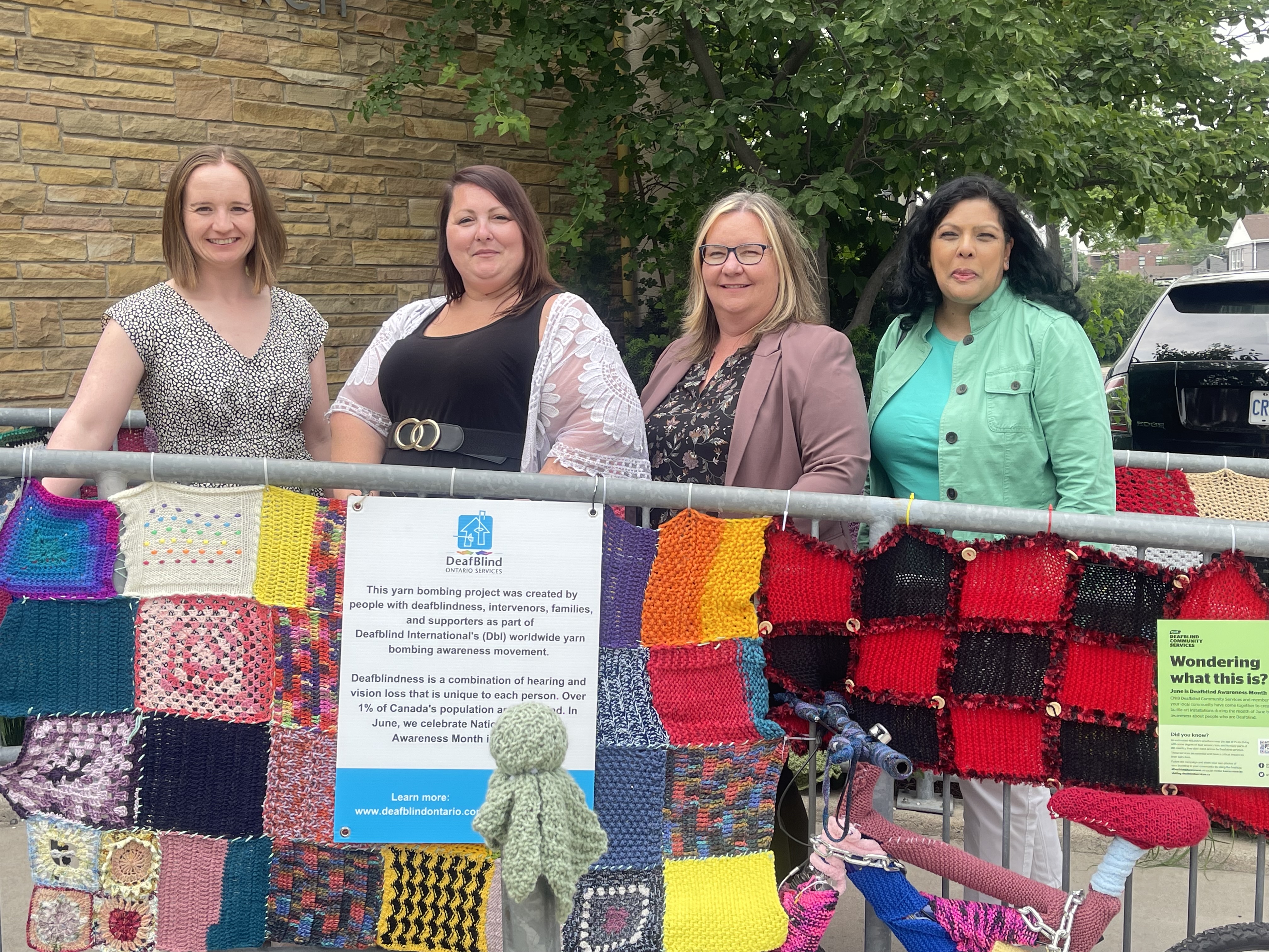 A group of people stand behind a yarn bombing display. there is a colourful yarn blanket on a fence in front of them 