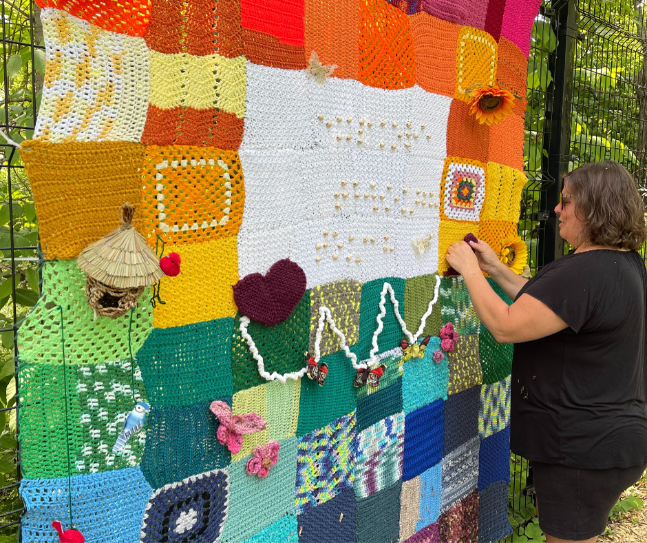 A woman places the finishing touches on a colourful yarn blanket that is hung against a fence. There are several tactile elements, and braille that can be felt. 