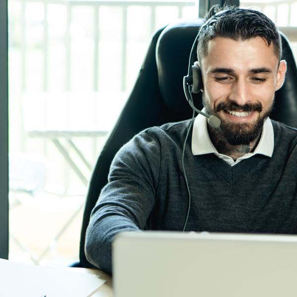 Un homme est assis devant un écran d’ordinateur. Il porte un casque d’écoute et sourit.