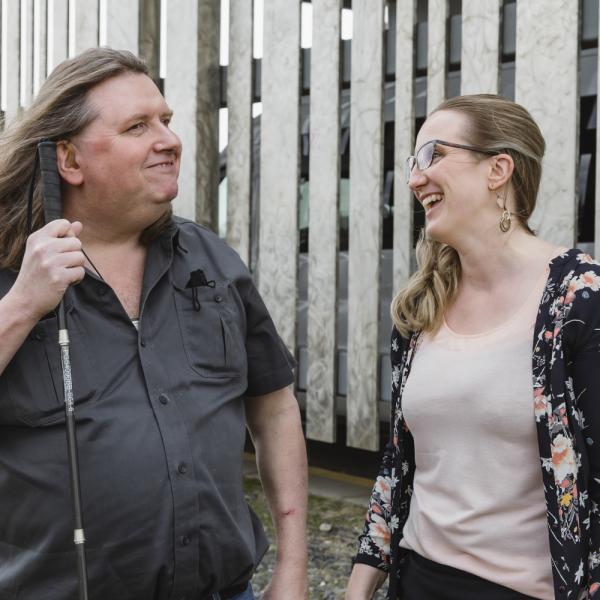 A DBCS client smiles while holding his cane. An intervenor stands beside him, with a big smile on her face.