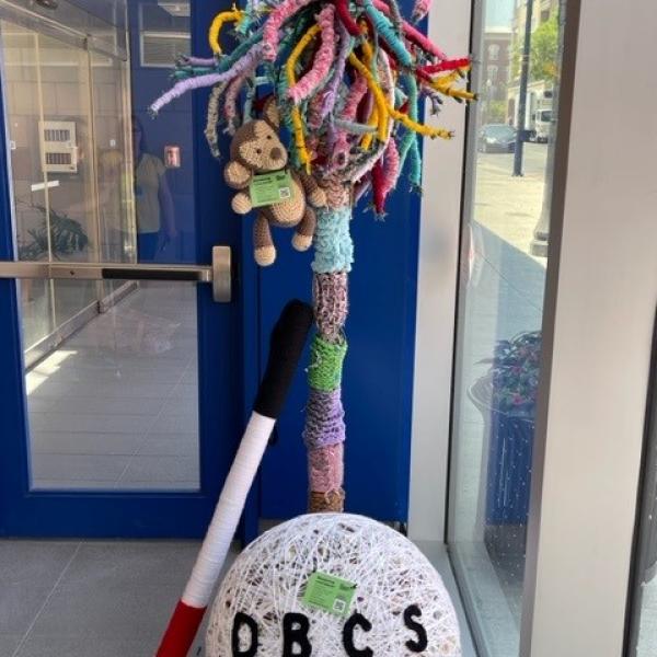 A giant yarn ball, with the text, DBCS stitched in english and braille on it. There is also a giant white cane made of yarn, and a colourful tree with a monkey in it. 