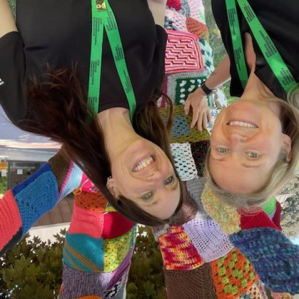 Two women stand in front of a tree covered in yarn. They are wearing green DBCS lanyards. 