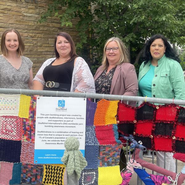 A group of people stand behind a yarn bombing display. there is a colourful yarn blanket on a fence in front of them 